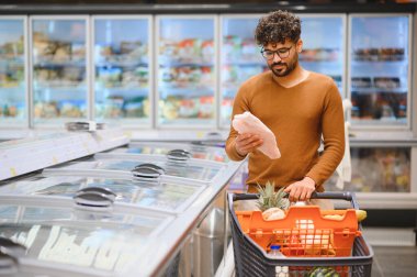 Genç adam market alışverişi yapıyor, modern bir süpermarketin soğutulmuş koridorunda donmuş balık paketini dikkatle inceliyor.