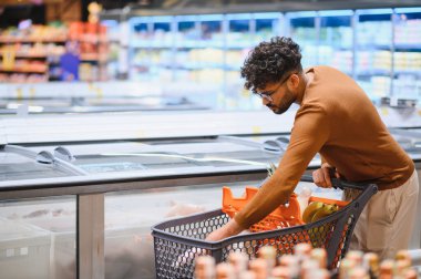 Genç adam market alışverişi yapıyor, buzdolabından dondurulmuş gıdalar seçiyor, alışveriş deneyiminin tadını çıkarıyor.