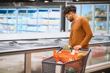 Müşteri market alışverişi yapıyor, alışveriş arabasını itiyor ve dondurulmuş yiyecekleri süpermarket buzdolabı bölümünden seçiyor.