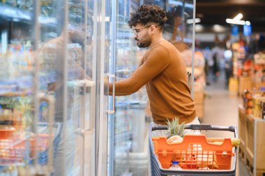 Genç Arap adam buzdolabının kapısını açıyor, markette dondurulmuş ürünleri seçiyor, alışveriş arabasını sağlıklı yiyeceklerle dolduruyor.