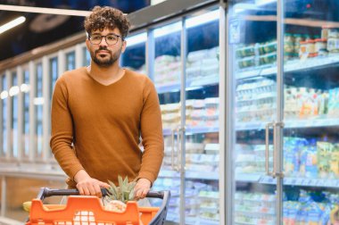 Genç bir adam süpermarkette alışveriş arabasını itiyor, dondurulmuş yiyecekleri ve taze sebzeleri seçerken soğuk koridorlarda geziniyor.