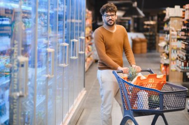 Marketin içinde alışveriş arabasını iten genç adam soğutulmuş koridorların yanında yürüyor.