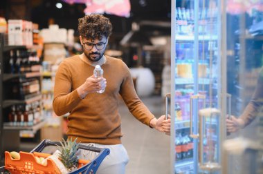 Genç Arap adam markette buzdolabı açarken bir şişe suyu kontrol ediyor, alışveriş arabasıyla alışveriş yapıyor.
