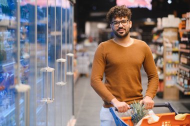 Genç Arap bir adam alışveriş arabasını iterken bir yandan da çeşitli yiyecek seçenekleriyle dolu, hareketli bir süpermarketten taze yiyecek seçiyor.