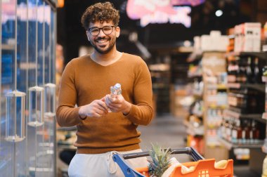 Müşteri süpermarkette bir şişe su seçiyor, markette içi yiyecek dolu bir alışveriş arabasını itip sağlıklı seçenekleri değerlendiriyor.