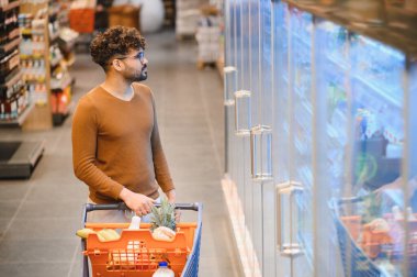 Genç adam alışveriş arabasını itiyor ve süpermarket koridorundaki cam vitrinden donmuş yiyecek seçiyor.