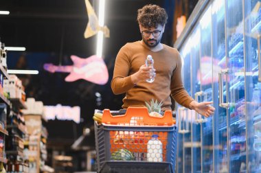 Müşteri bir süpermarket koridorunda dolaşırken bir şişe etiketini inceliyor, market poşetleri ve taze ürünlerle dolu dolu bir arabayı itiyor.