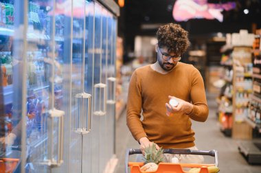 Bir süpermarketin market koridorlarında dolaşırken ürün malzemelerini inceleyen genç adam sağlıklı beslenmek için bilgilendirilmiş seçimler yapıyor.