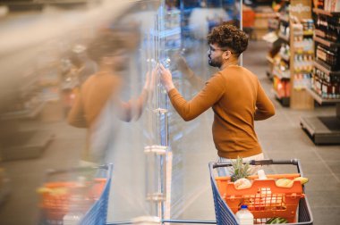 Arap bir adam markette buzdolabının kapısını açarken market alışverişini seçiyor, tipik bir alışveriş deneyimi yaşıyor.