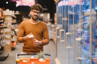Süpermarket buzdolabı koridorunda alışveriş yaparken market ürünlerine etiket okuyan genç adam.