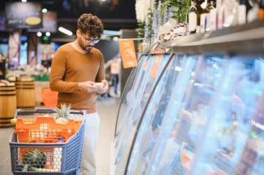 Genç Arap adam alışveriş arabasını itiyor ve süpermarketteki soğutulmuş ürünlerin etiketlerini kontrol ediyor.