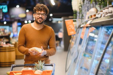Genç adam elinde taze bir somun ekmek tutarken markette alışveriş yapıyor, gülümsüyor ve bu deneyimin tadını çıkarıyor.