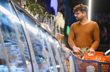 Müşteri alışveriş arabasını iterken modern bir süpermarketin buzdolabı bölümünden taze sebze seçiyor.