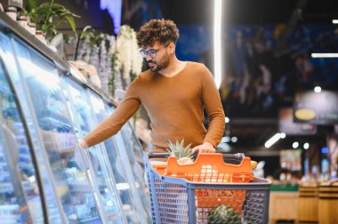 Müşteri, süpermarketin soğutuculu bölümünden taze sebze seçerken parlak koridorlarda bir alışveriş arabasını itiyor.