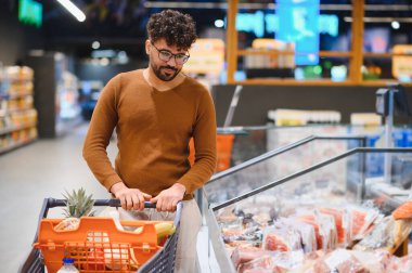 Genç Arap bir adam alışveriş arabasını iterken iyi organize edilmiş bir süpermarket buzdolabından taze et ürünleri seçiyor.