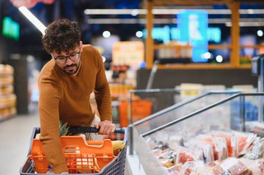 Genç Arap bir adam alışveriş arabasını iterken markette taze yiyecekler seçiyor, sağlıklı bir yaşam tarzını somutlaştırıyor.