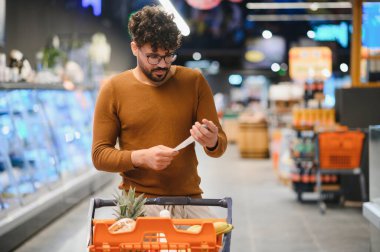 Bakkal fişini kontrol eden odaklı Arap müşteri süpermarket koridorunda alışveriş arabasını iterken, harcamaları yönetirken ve doğru faturaları garantilerken