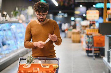 Genç bir Arap süpermarkette alışveriş arabasını iterken market fişini kontrol ediyor. Masrafları ve bütçeyi yönetmeye odaklanıyor.