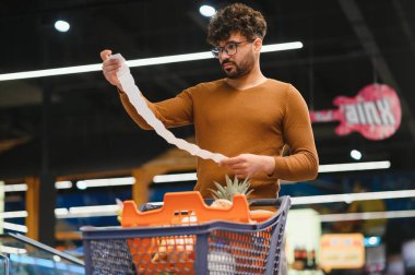 Alışverişci market faturasını inceliyor, satın alma miktarına şaşırmış, süpermarkette kişisel finansman yönetiyor.