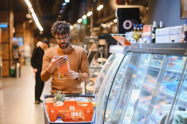 Genç adam bir markette alışveriş arabasını iterken yiyecek paketindeki malzemeleri dikkatlice kontrol ediyor.