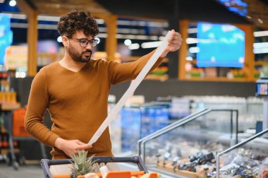 Müşteri, süpermarkette alışveriş yaparken kapsamlı bir market fişini kontrol ediyor, artan harcamalar ve maliyetler konusunda şaşırmış ve endişeli görünüyor