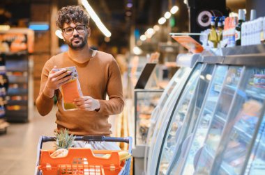 Süpermarkette alışveriş yapmayı seçen, vakumlu somon etiketi okuyan, alışveriş arabasını iten genç bir adam.