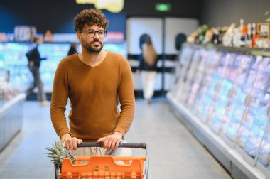 Genç bir Arap süpermarkette alışveriş arabasını itiyor, dondurulmuş raflardan yiyecek seçiyor.