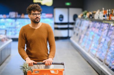Genç Arap bir adam alışveriş arabasını iterken parlak ışıklı bir süpermarkette taze yiyecekler seçiyor ve koridorlarda rahatça geziniyor.