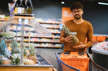 Genç bir Arap süpermarkette ananas seçiyor, alışveriş arabası tutuyor ve market alışverişi yapıyor.