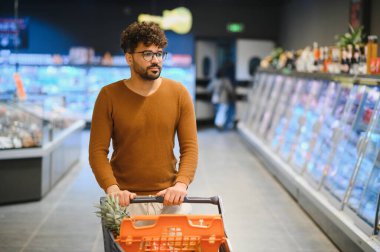 Genç bir adam market koridorunda alışveriş arabasını itiyor, taze yiyecek seçiyor ve alışveriş deneyiminin tadını çıkarıyor.