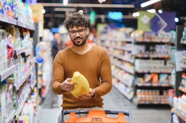 Müşteri süpermarkette deterjan seçiyor, şişeyi tutuyor, etiketi okuyor, market alışverişi yapıyor.