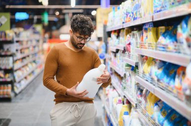 Genç Arap adam süpermarkette alışveriş yaparken temizlik ürününün etiketini dikkatlice okuyor.