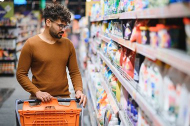 Genç Arap bir adam alışveriş arabasını iterken parlak ve modern bir süpermarket koridorundan taze yiyecek seçiyor.