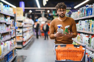 Müşteri, süpermarket koridorunda alışveriş yaparken iki farklı temizlik ürününü karşılaştırıyor, etiketleri dikkatlice okuyor ve bir seçim yapıyor.
