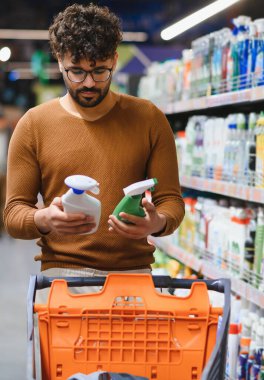 Markette alışveriş yaparken temizlik ürünleri seçen, raflardaki seçenekleri karşılaştıran ve ev ihtiyaçları için etiketleri inceleyen bir adam.