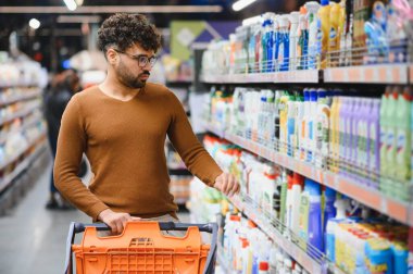 Genç adam parlak ışıklı bir süpermarkette temizlik ürünlerini dikkatlice seçiyor, bir alışveriş arabasını itiyor ve koridorlarda geziniyor.