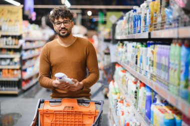 Genç adam süpermarkette temizlik ürünleri alıyor, elinde bir şişe tutuyor ve alışveriş arabasını itiyor.