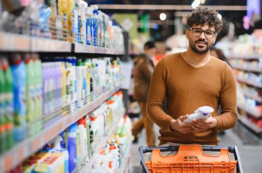 Genç Arap adam, markette elinde alışveriş arabasıyla temizlik ürünleri etiketini seçiyor.