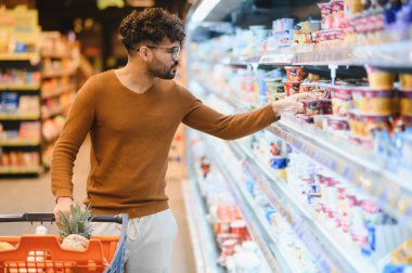 Genç yetişkin marketin buzdolabı bölümünde yoğurt seçiyor, meyve dolu bir alışveriş arabasını itiyor.