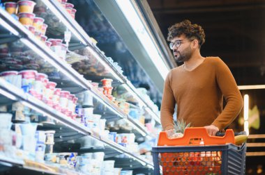 Genç Arap adam alışveriş arabasını iterken süpermarketin soğutulmuş bölümündeki yiyecekleri seçiyor.