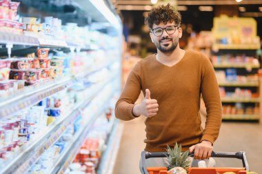 Bir süpermarkette alışveriş arabasını iterken mutlu müşteri el kol hareketi yapıyor, alışverişlerinden memnun.