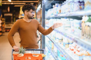Genç Arap adam alışveriş arabasını itiyor ve süpermarketin soğutulmuş bölümünden süt şişesini seçiyor.