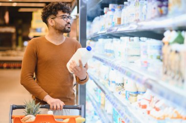 Genç Arap adam bir süpermarketin mandıra bölümünde bir şişe süt seçerken bir alışveriş arabasıyla alışveriş yapıyor.