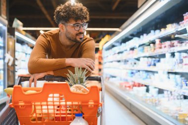 Genç Arap adam gözlük takıyor, alışveriş arabasına yaslanıyor ve canlı bir süpermarket koridorunda düşünceli bir şekilde erzak seçiyor.