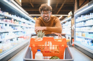 Müşteri alışveriş arabasına yaslanıyor. Taze yiyeceklerle dolu. Hareketli bir süpermarketin canlı atmosferinin tadını çıkarıyor.