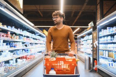 Genç bir Arap süpermarketin koridorlarında alışveriş arabasını itiyor ve taze ürünleri seçiyor.