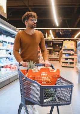 Müşteri parlak bir market koridorunda alışveriş arabasını itiyor, canlı vitrinlere bakarken taze ürünleri seçiyor.