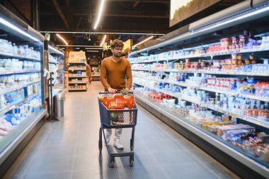 Müşteri süpermarkette alışveriş arabası itiyor, koridorlardan çeşitli gıda ürünleriyle dolu taze yiyecekler seçiyor.