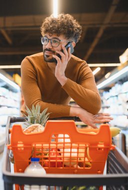 Sakallı bir adam markette alışveriş arabasının üzerine yaslanırken bir telefon görüşmesi yapıyor.