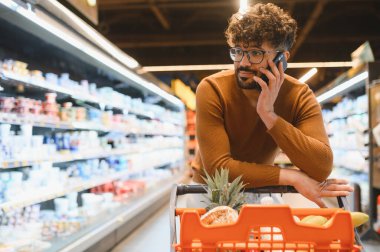 Market koridorunda alışveriş arabasına yaslanarak akıllı telefondan konuşan, bakkaliyeyi seçen bir müşteri.